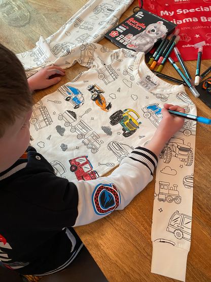 Child colouring vehicle-themed pyjamas with included fabric pens on a wooden table.