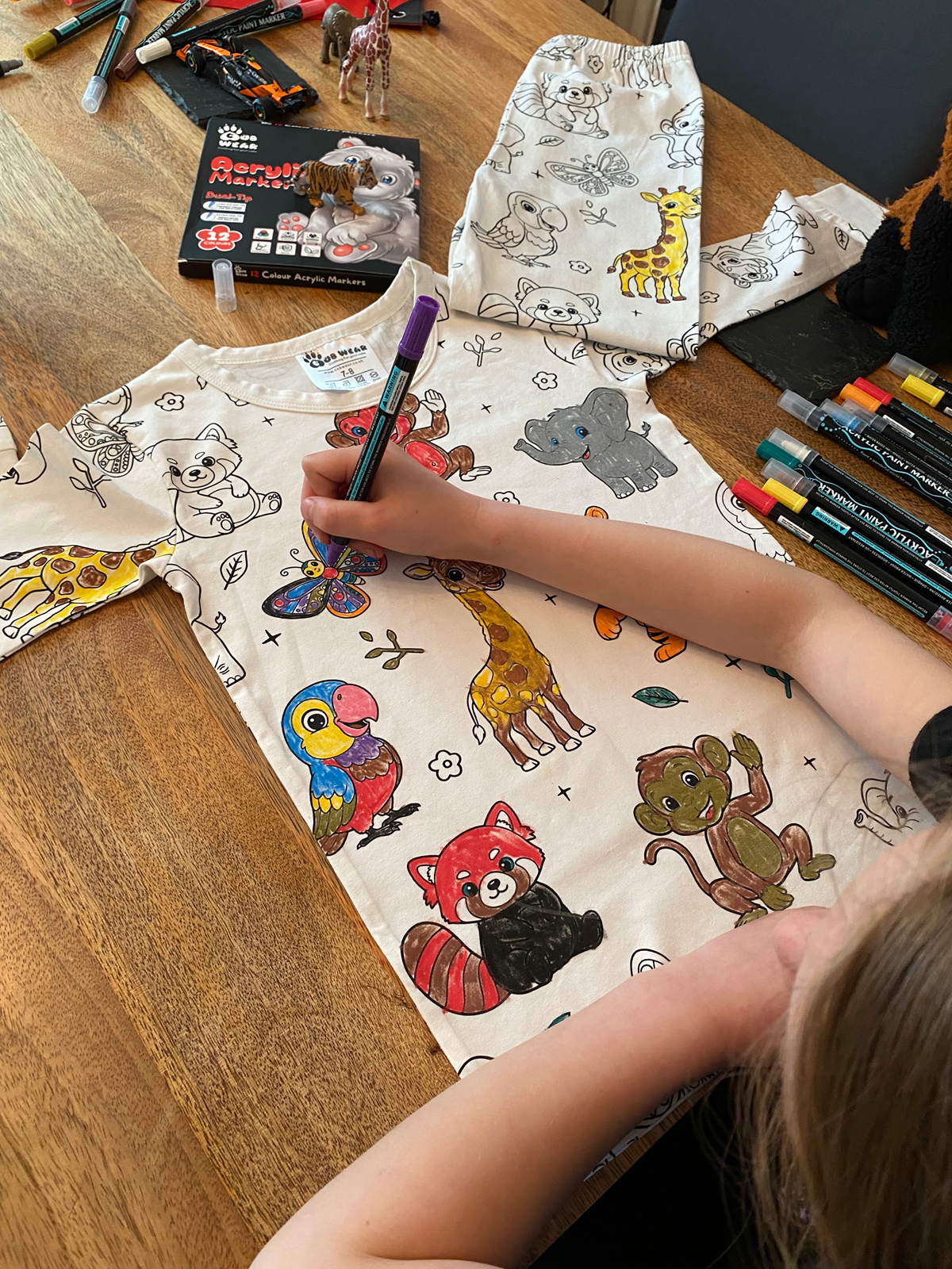 Child colouring animal-themed pyjamas using included fabric pens on a wooden table.