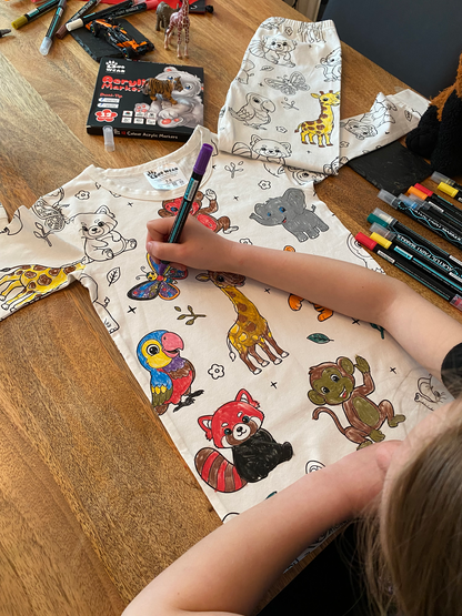 Child colouring animal-themed pyjamas using included fabric pens on a wooden table.